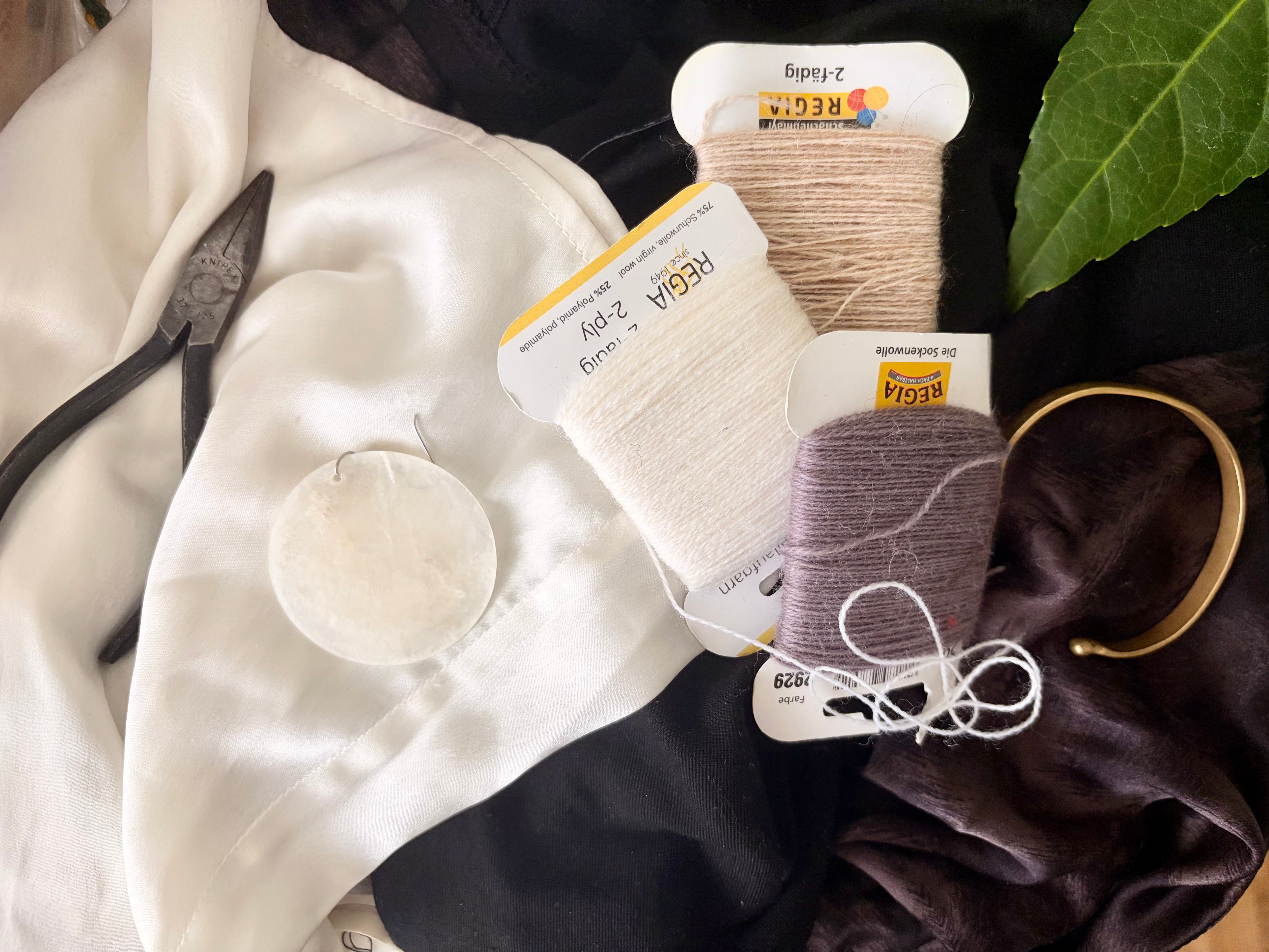 Flat-lay of sewing materials: silk fabric, thread spools, a pearl button and a gold bracelet